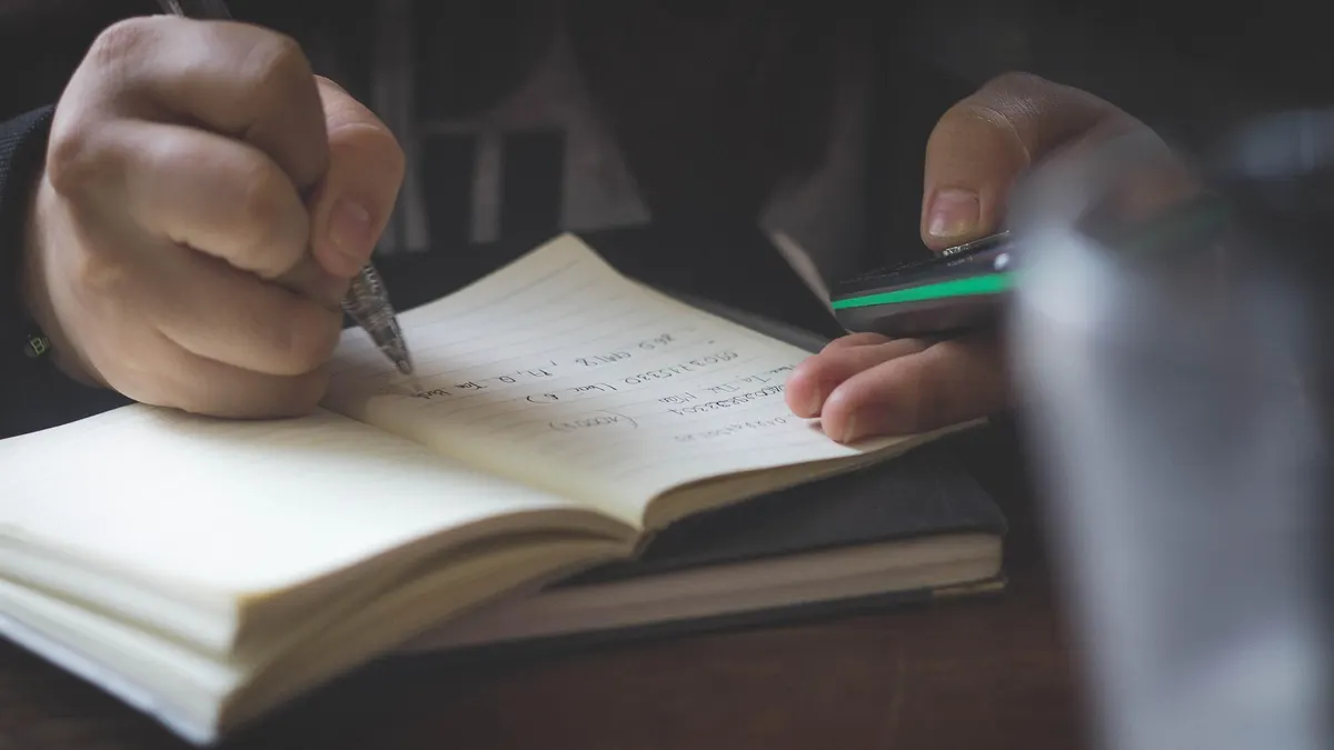 Hand taking notes in a small notebook next to a glass of water